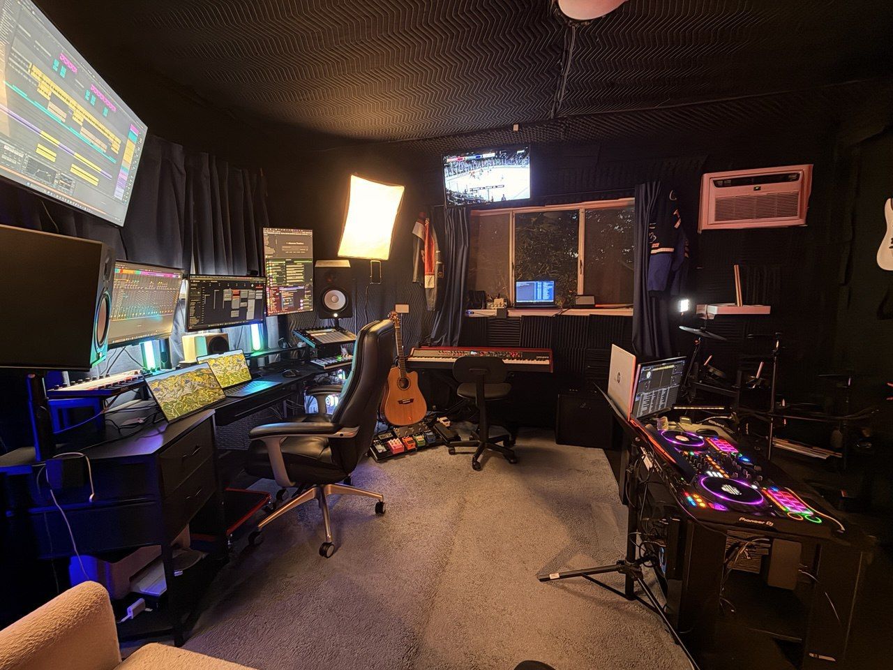 Behind the desk production setup with monitors, DJ controller, guitar, and keyboard