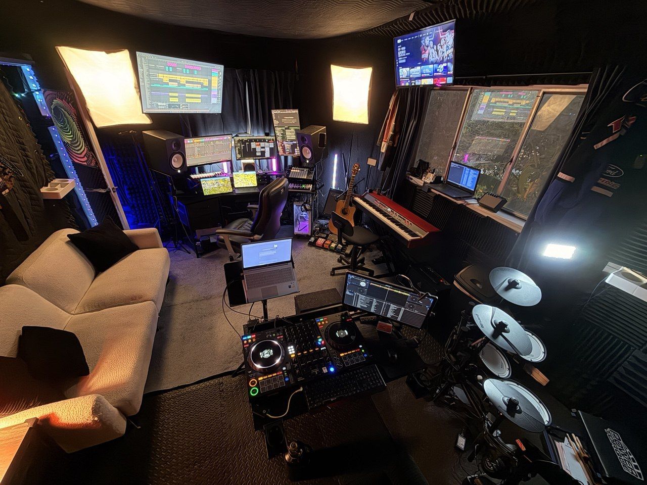 Elevated wide shot — full studio with DAW, DJ deck, drums, couch, multiple screens