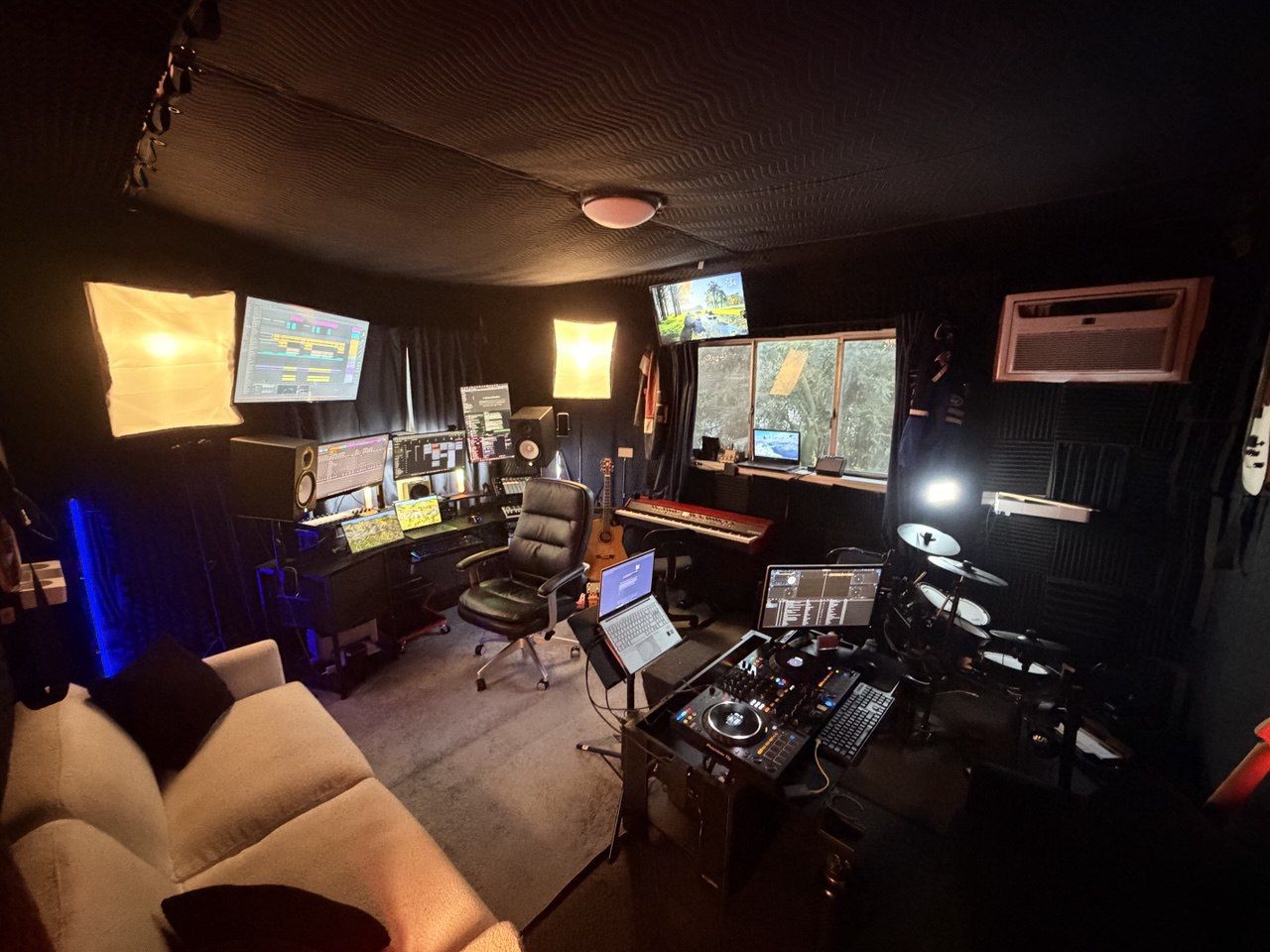 Clean studio view from couch with DAW monitors, lights, DJ controller, and drums